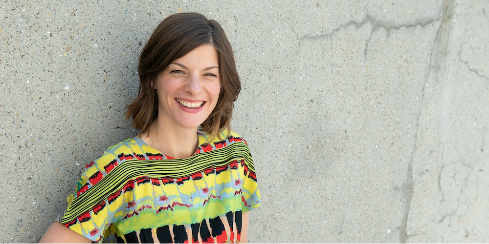 Associate Professor Desiree Fields, smiling, wearing a colorfully patterned blouse in front of grey concrete background.