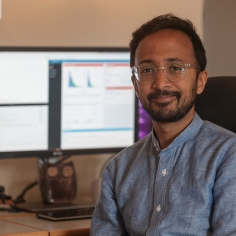 Ajay Pillarisetti setting at desk with computer monitor and microphone in background