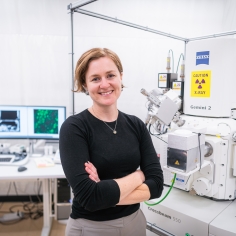 Assistant Professor Bronwyn Lucas stands with arms crossed next to a Zeiss Crossbeam cryo-FIBSEM.