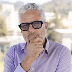 person with short gray hair and black rim glasses holding chin