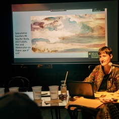 Image of a white disabled woman in her forties. She is speaking into a microphone giving a presentation. Image is of Sunaura Taylor. 