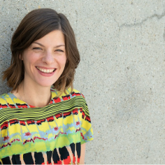 Associate Professor Desiree Fields, smiling, wearing a colorfully patterned blouse in front of grey concrete background.