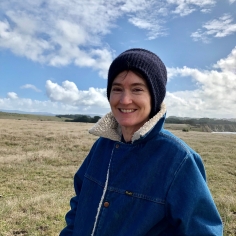 Steph wearing a denim coat with collar upturned, and with a big blue sky and white puffy clouds in the distance.
