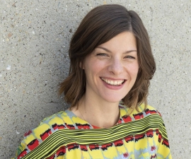 Associate professor Desiree Fields, smiling, wearing a colorfully patterned blouse in front of grey concrete background.