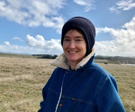 Steph wearing a denim coat with collar upturned, and with a big blue sky and white puffy clouds in the distance.