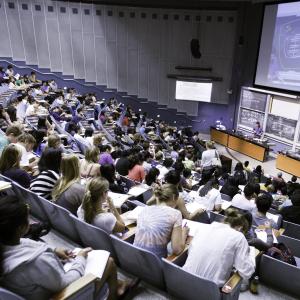 lecture hall from behind showing students backs and a faculty at the front of the hall with overhead screen of diagram 