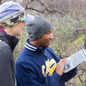 Two people in nature looking at a laptop held by one of them.