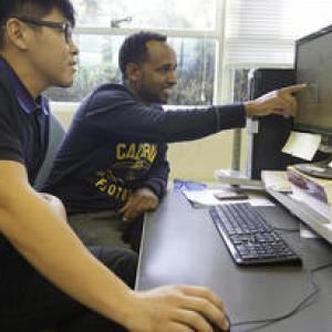 Two people sitting at a computer monitor, one is pointing at the screen.