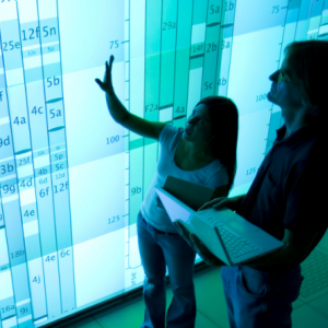 2 people standing in front of a digital board of data references