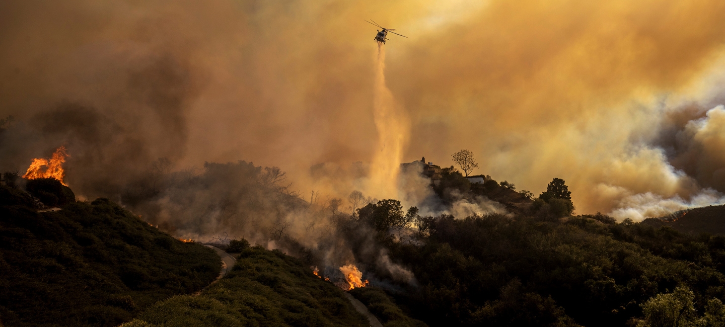 A helicopter in the center frame drops water on a fire as yellowish smoke clouds the sky. Ethan Swope/AP