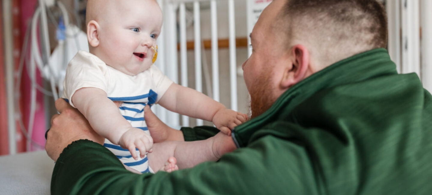 a man in a green hoodie holding a smiling baby with both arms