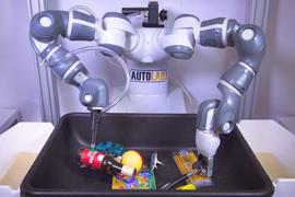 A grey robot with a suction gripper on one arm and a claw gripper on the other is poised above a container of objects.
