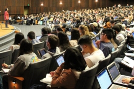 A photo of a large auditorium filled with students all listening to a single speaker on stage.