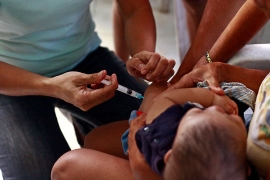 A photo shows a pair of arms and hands holding a syringe, injecting a small child