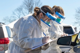 Two National Guard health officers stand outside next to a car wearing personal protective suits and face shields, and writing on clipboards