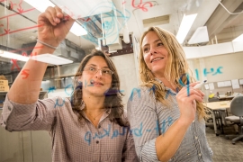 A photo of two women writing mathematical equations on a piece of glass