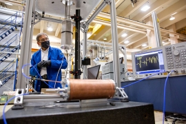 A faculty member wearing a face mask and a blue lab coat stands in a laboratory. He stands behind a copper tube, some silver scaffolding, and a flashing screen.
