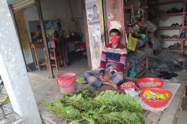 A photo of a Bangladeshi shop keeper wearing a red cloth face mask, while sitting outside behind a selection of produce.