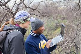 Two students look at a laptop while standing outside in a wooded area