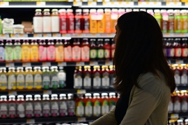 A women shopping in a grocery store shopping with a cart and a wall of beverages in the background.