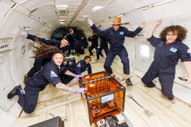 A group of people wearing blue jumpsuits surround a clear orange box that is attached to the floor in the cabin of an aircraft. They are smiling and floating in the air because they are experiencing zero gravity.