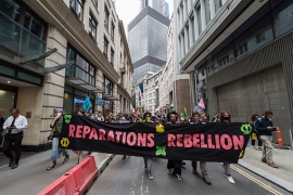 A photo shows protestors marching down a city street holding a large sign that reads "Reparations, Rebellion," in large, pink on black letters