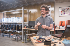 A photo of a student standing at at table, smiling, with a bank of 3D printers in the background.