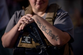 A smiling person with a large rifle and "We the People" tattooed on her arm