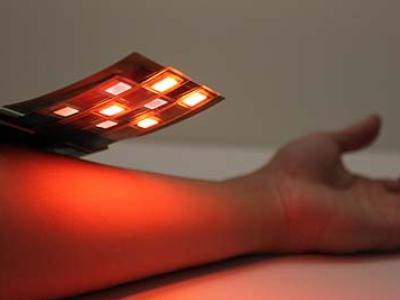 A thin, flexible electronic board, lit-up with red and infrared lights, and approximately three inches wide by three inches wide, is held above a person's forearm.