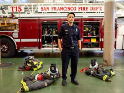 A photo of Maiko Bristow, a women firefighter with the San Francisco Fire Department. Maiko is wearing a black uniform and her turn out gear sits on the ground around her. Behind her is a fire truck with the words "San Francisco Fire"