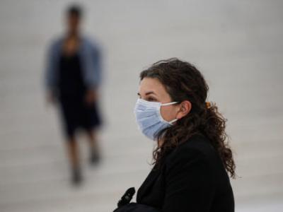 A woman stands on a street wearing a light blue surgical mask.