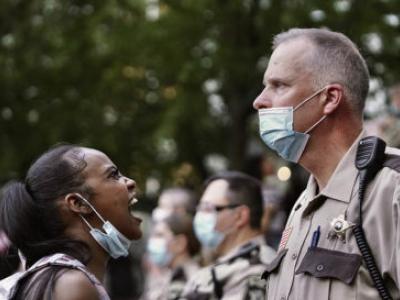 a black woman with a mask at her chin yells at a white male police officer wearing a mask and staring into the distance