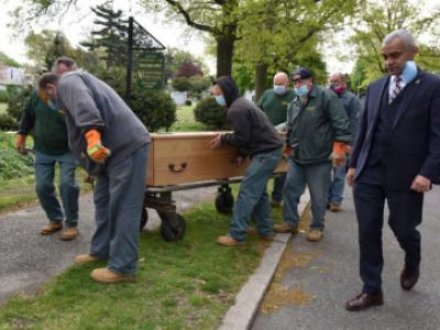 a man in a suit walks along with a wooden casket being rolled by workers in masks 