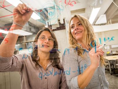 A photo of two women writing mathematical equations on a piece of glass