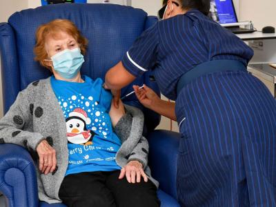 a woman wearing a mask sits in a blue armchair and gets a shot in her arm