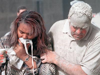 a woman with her face covered against fumes is supported by an ash-covered man near the site of the World Trade Center collapse