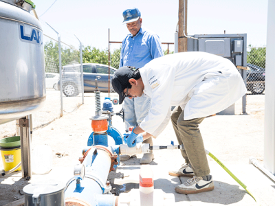 On a bright sunny day, a person wearing a white lab coat and a black baseball cap leans over to collect a water sample from an outdoor well. A second person, wearing a blue denim shirt, stands behind them.
