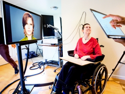 Research participant, sitting in a wheelchair, is connected by wires to a computer and monitor that displays an avatar. A research coordinator can be seen walking behind the monitor, and in the foreground, someone is holding a laptop computer.