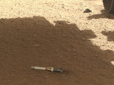 a small white and black tube sits on pebbly red ground in the shade of the Insight lander on Mars
