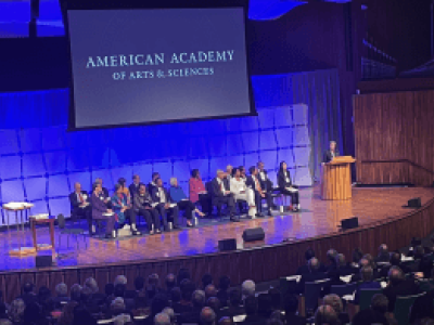 Multiple people, presumably scholars, sitting in chairs on the stage of an auditorium, overlooking an audience. One person is standing at a podium on the stage. A projected image behind the people on the stage reads "American Academy of Arts & Sciences." 