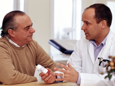 an older man, at left, confers in a medical office with a doctor (at right) who is holding out a prescription vial.