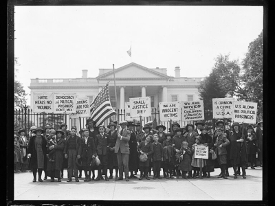 In a black and white photograph, a group of people, including children, stand in front of the White House holding signs that say "shall justice die?", amongst other posters