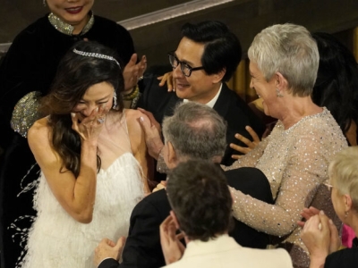 Michelle Yeoh stands with a hand to her mouth as Jamie Lee Curtis and Ke Huy Quan congratulate her.