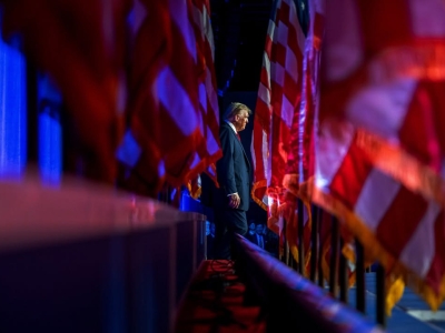 With a row of American flags on the left and right side of the frame, Donald Trump walks in the distance while supporters cheer