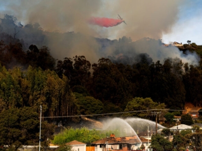 Smoke emerges from forested hills. In the foreground, two fire trucks aim streams of water at a vulnerable home. Overhead, an aircraft releases a plume of red flame retardant chemicals.