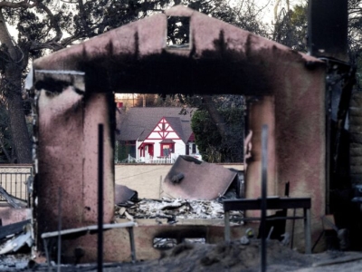 A charred building, surrounded by debris and ash with a view through the structure toward a white-painted home that is intact and undamaged by fire.