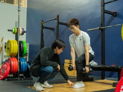 a trainer wearing dark clothing kneels and directs a student who is doing a squat with dumbbells