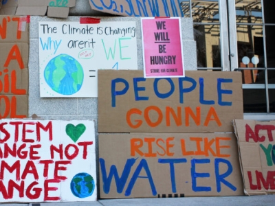 A collection of posters about climate change and the need to take policy action are posted on the steps of a public building. The posters have various messages such as "don't be a fossil fool."