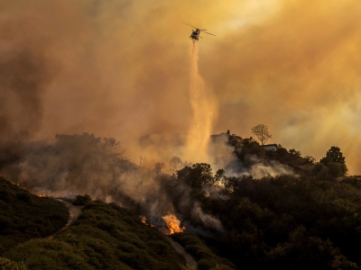 A helicopter in the center frame drops water on a fire as yellowish smoke clouds the sky. Ethan Swope/AP