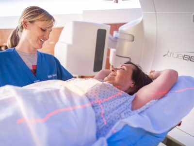 A woman in a medical gown lies on her back inside a diagnostic machine, smiling at a technician preparing to take an image.